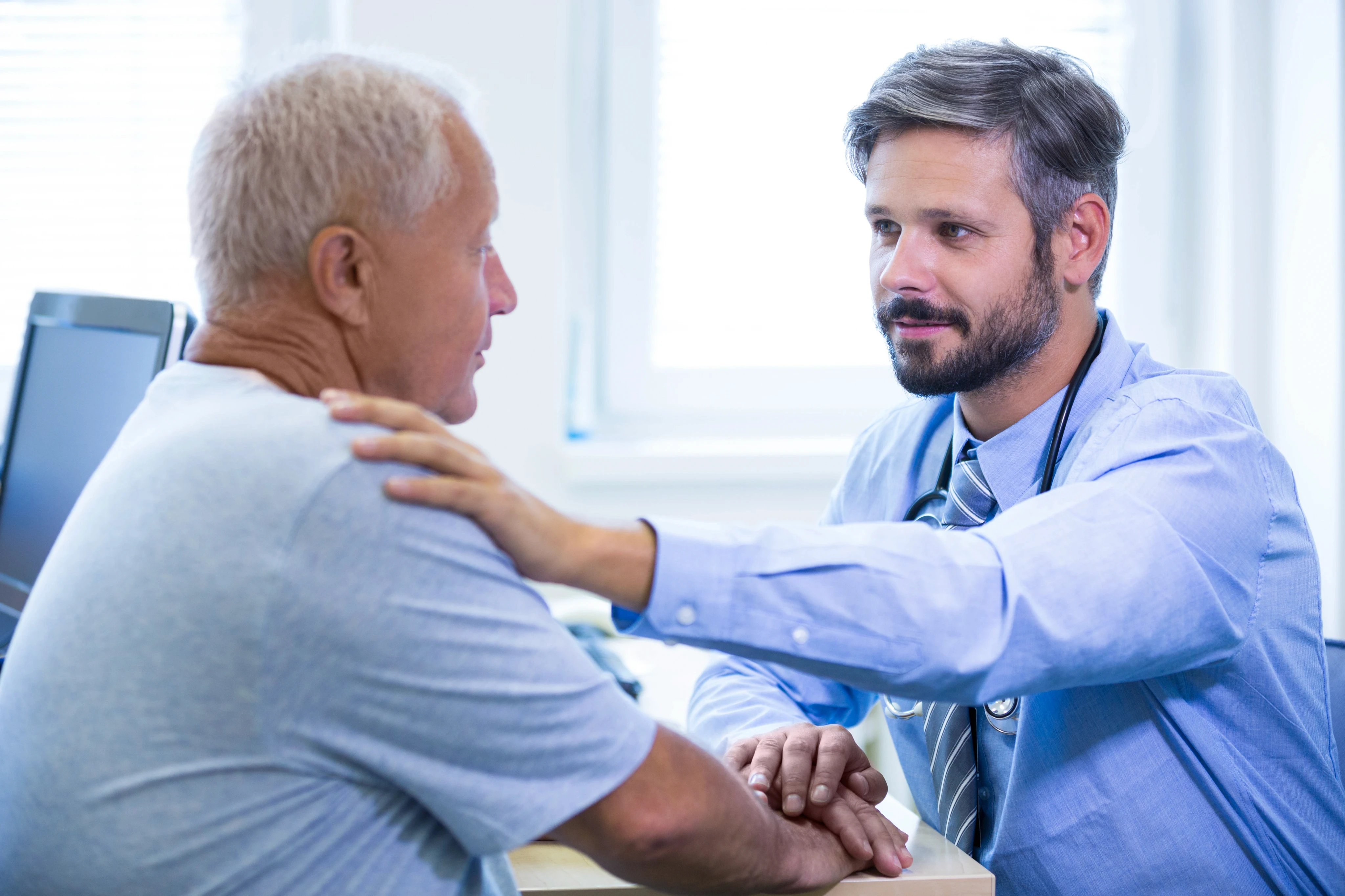 A doctor in a blue shirt, with a stethoscope, compassionately places a hand on an elderly man's shoulder during a consultation in a bright office.