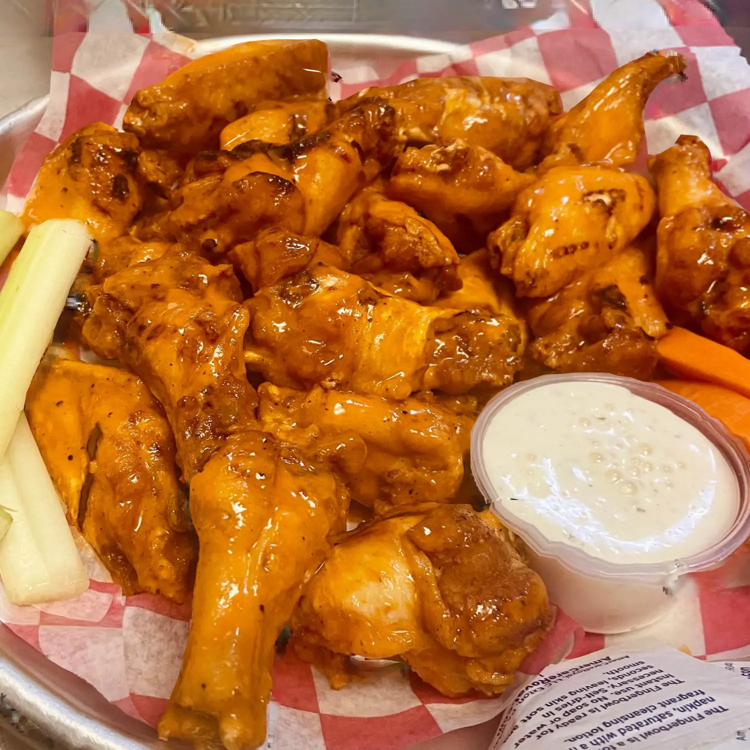 A platter of glazed chicken wings is served with celery sticks, carrot slices, and a side of creamy dipping sauce on a checkered paper-lined tray, exemplifying savory dining options in Lakewood Ranch, FL.