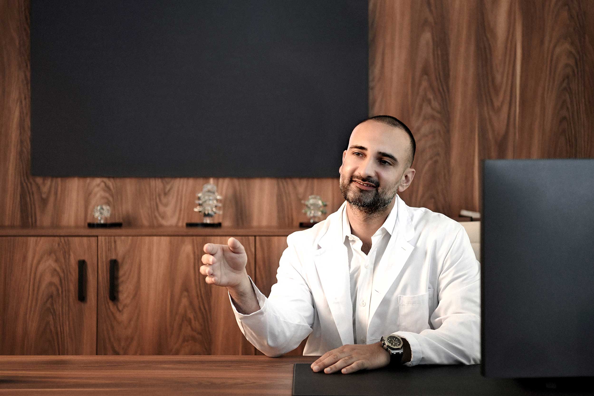 Dr. med. Sina Havakeshian im Beratungsgespräch in der Praxis SpinaMed, vor einer Holzvertäfelung mit kleinen Wirbelsäulenmodellen im Hintergrund.