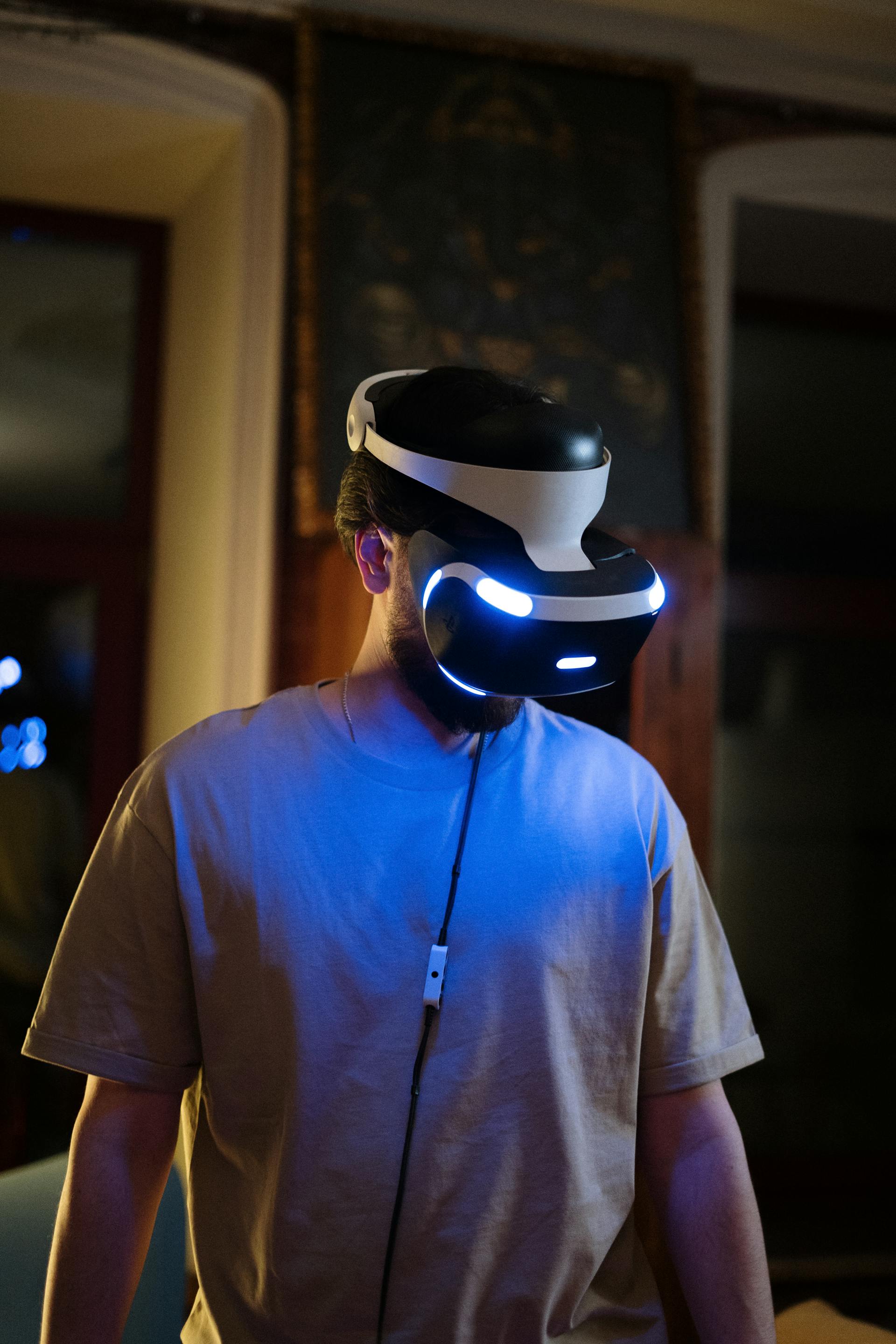 A person is wearing a virtual reality (VR) headset with glowing blue lights in a dimly lit indoor setting.
