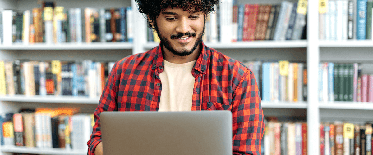 A student smiling while using a laptop in a library, representing the process of adapting traditional lesson plans for video.