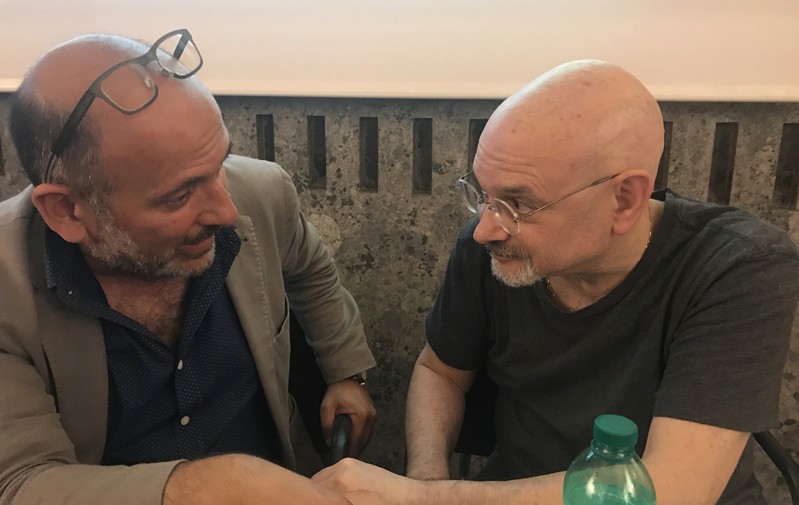 A photo of two men sitting at a desk and talking to each other.