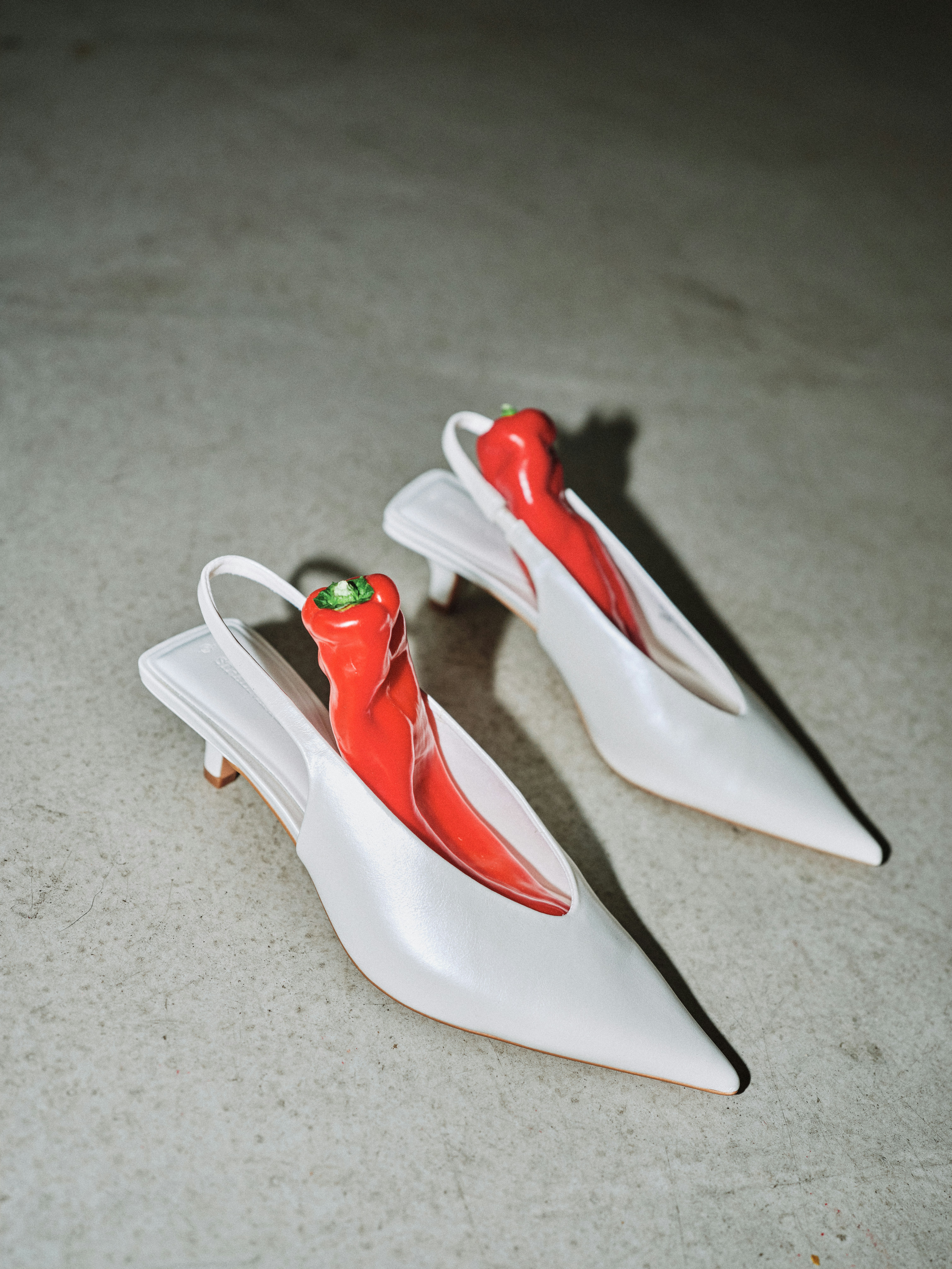 Surreal fashion still life of two white pointed kitten heel slingback shoes on a concrete floor, each containing a red pepper in place of a foot