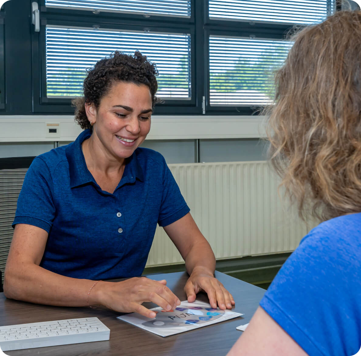A woman in a blue polo shirt sits at a desk, smiling and using a pen to explain something to another person, with a document and computer keyboard visible, suggesting a collaborative office environment.
