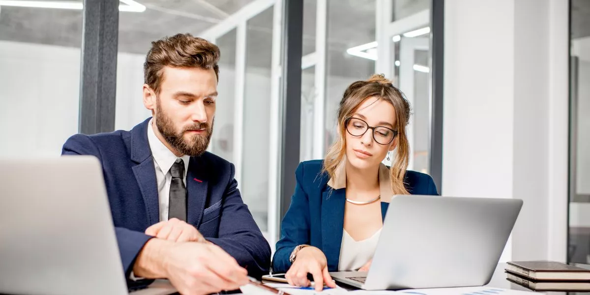 A man stands while two people sit at a table in an office setting, smiling and engaged in conversation. The mood is friendly and collaborative.