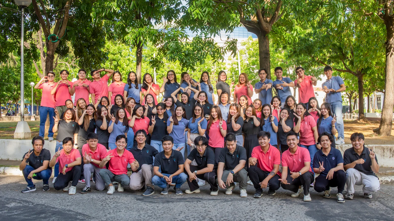 A large group of employees pose outdoors in coordinated shirts, smiling and making cheerful hand gestures under leafy trees.
