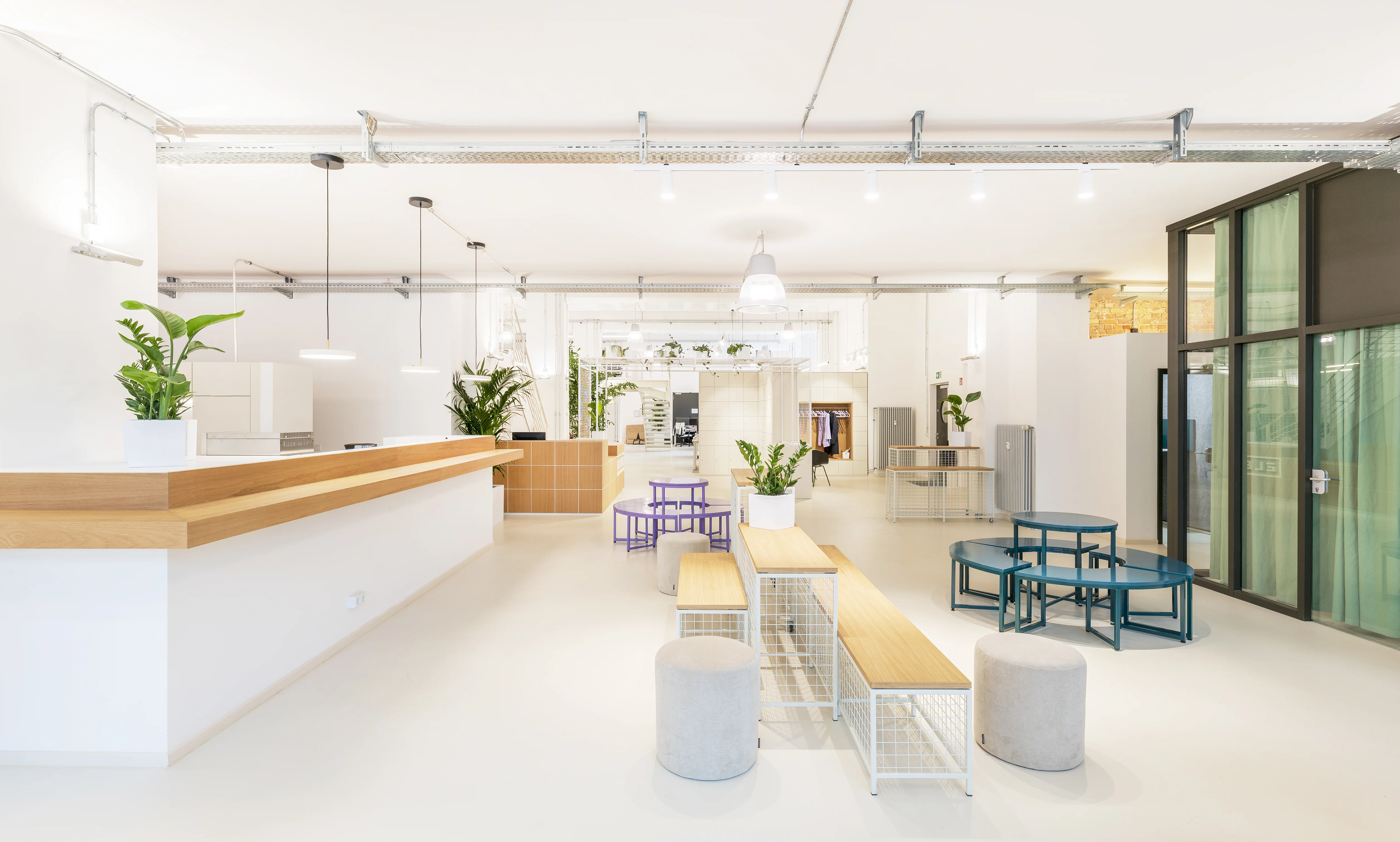 Open ground floor at ELEMENT with a long white counter with oak top, wooden benches on white mesh bases, grey poufs, petrol-blue and purple stool groups and an industrial glazed facade.