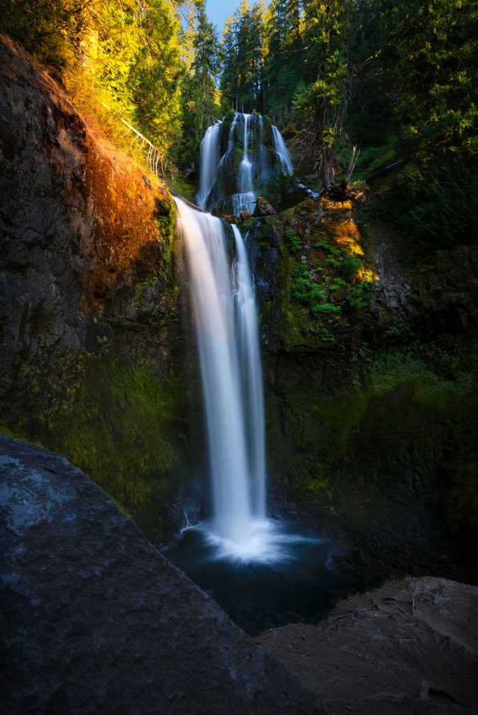 A waterfall into a body of water in a forest with sunlight peaking through the trees