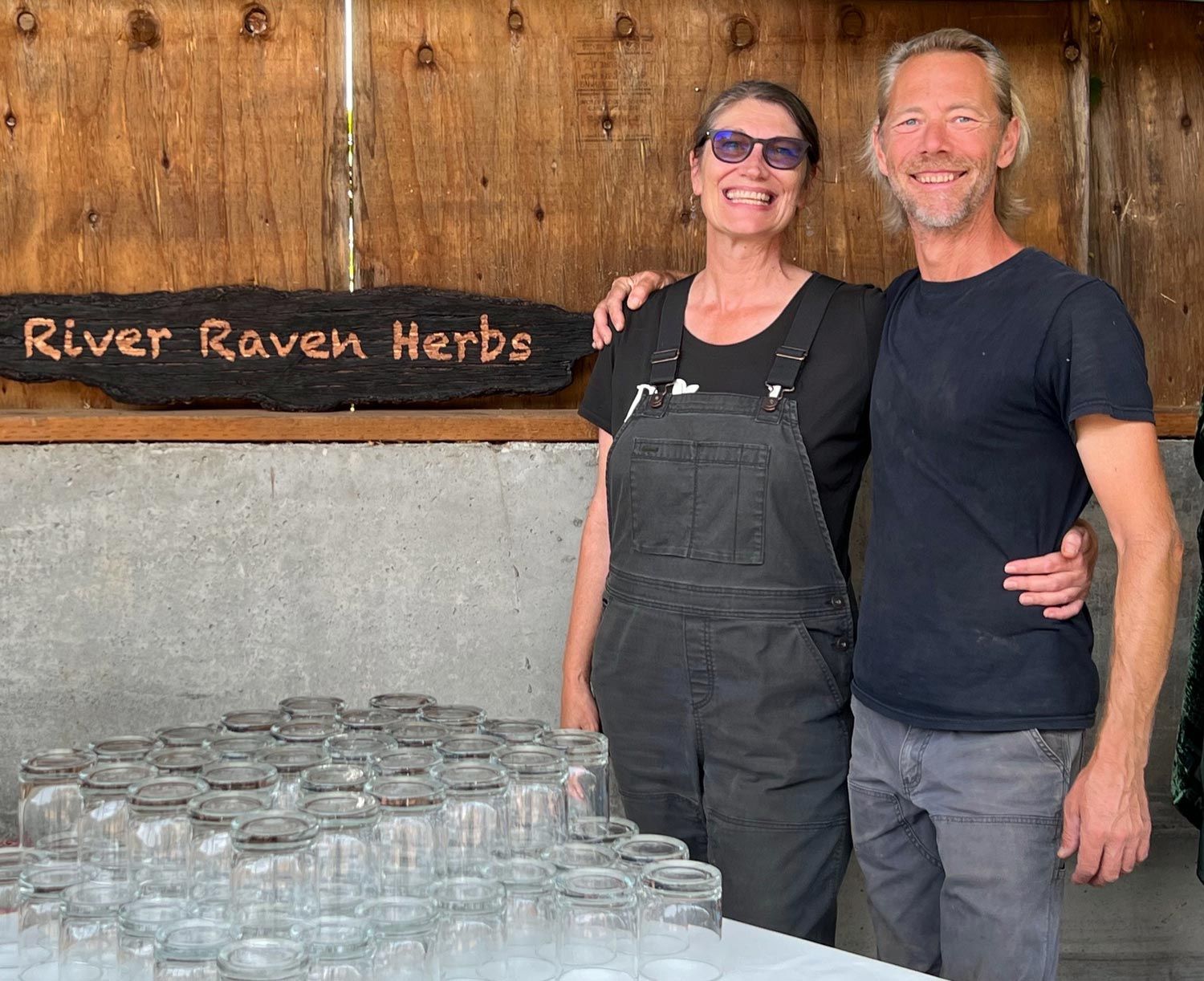 The owners of River Raven Herbs standing together in their workspace surrounded by jars and equipment.