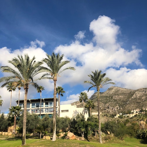 A modern building surrounded by palm trees and greenery, with mountains in the background under a partly cloudy sky.