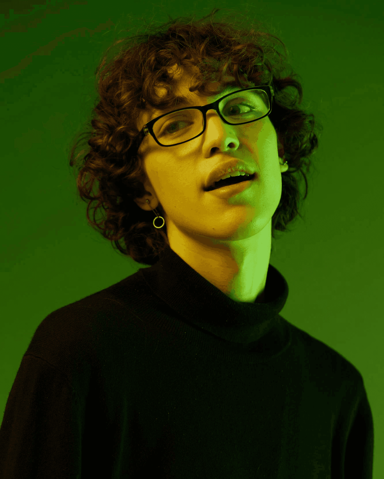Close-up of a young man with curly hair wearing black-rimmed glasses and a turtleneck, illuminated by vibrant green ambient lighting.
