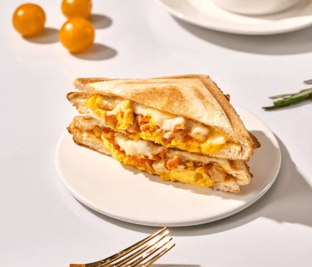 A delicious grilled cheese sandwich, cut in half, revealing melted cheese and scrambled egg products served on a white.