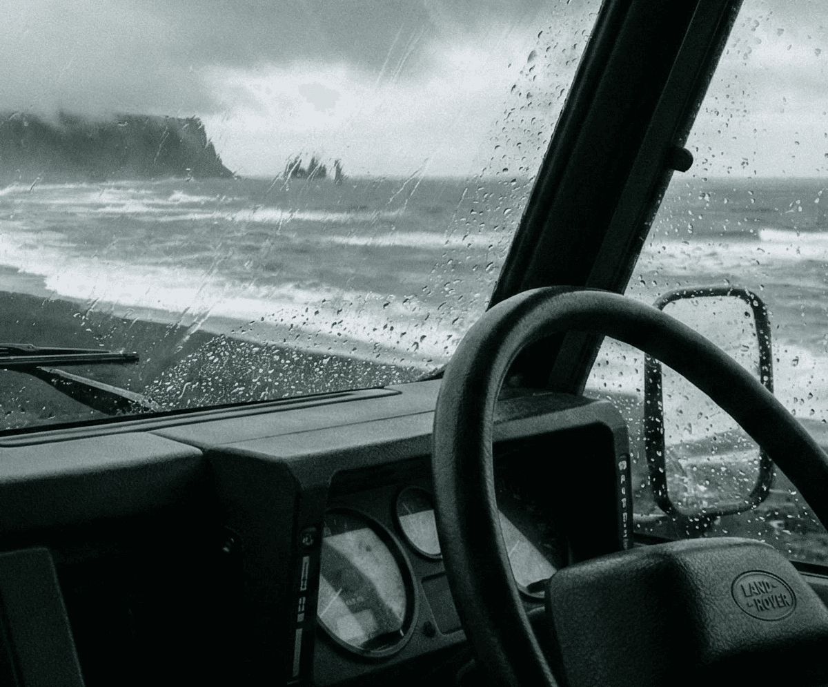 A view from the driver's seat looking through a rain-streaked windshield at another vehicle driving along a black sand beach near the ocean.