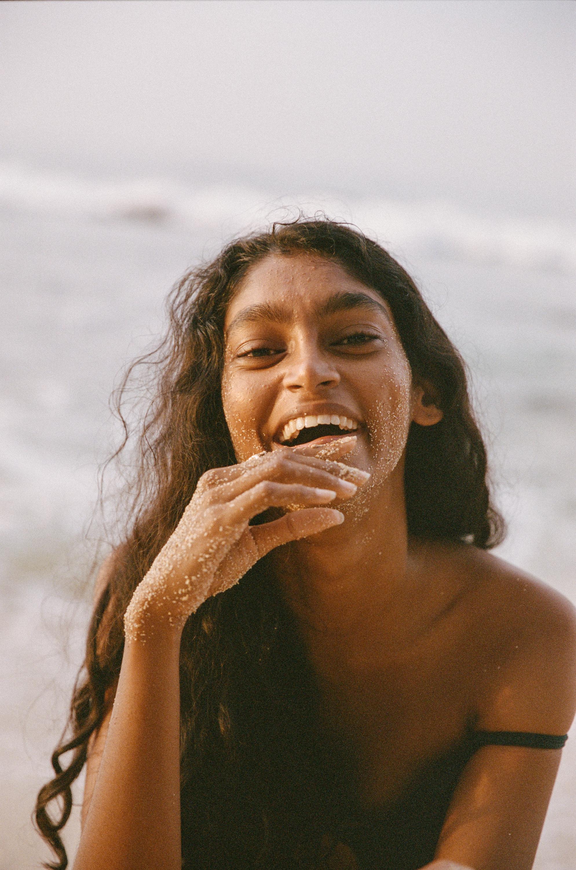 Woman smiling on the beach with sand on her skin and ocean waves in the background.