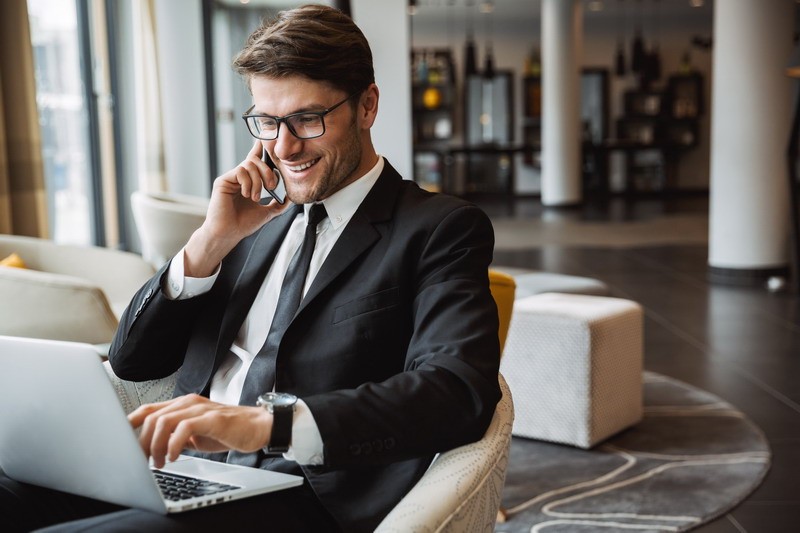 Photo of happy young businessman using laptop computer and talking on smartphone in hotel hall