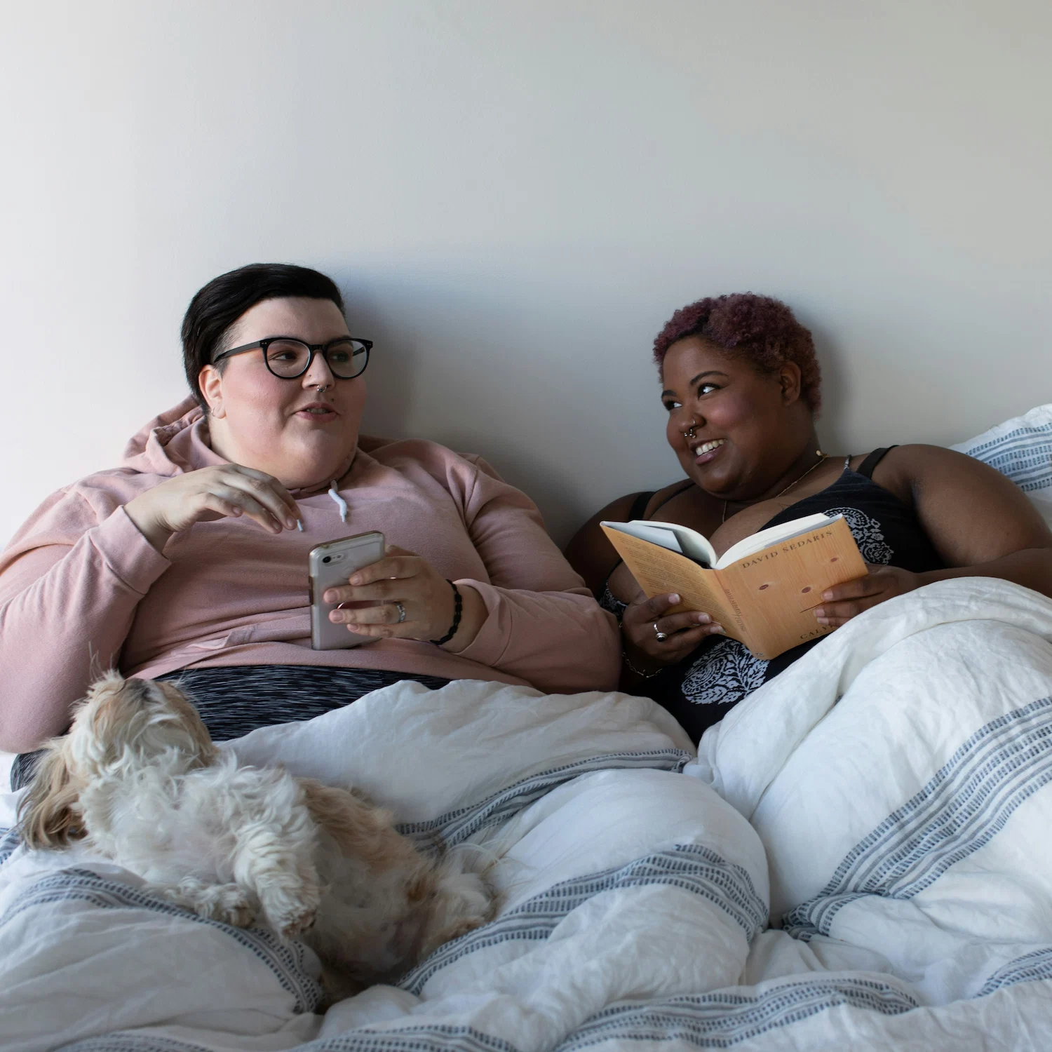 Happy lesbian couple relaxing in bed together with a book and phone, representing support, connection, and inclusivity in lesbian affirming therapy NYC.