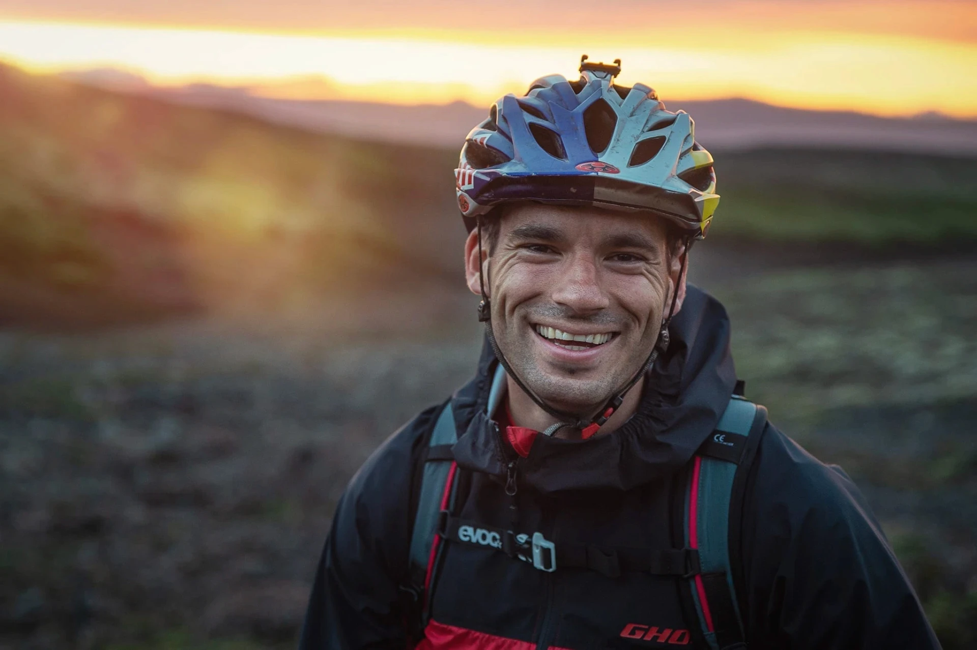 Smiling cyclist in a helmet and backpack with sunset light behind.