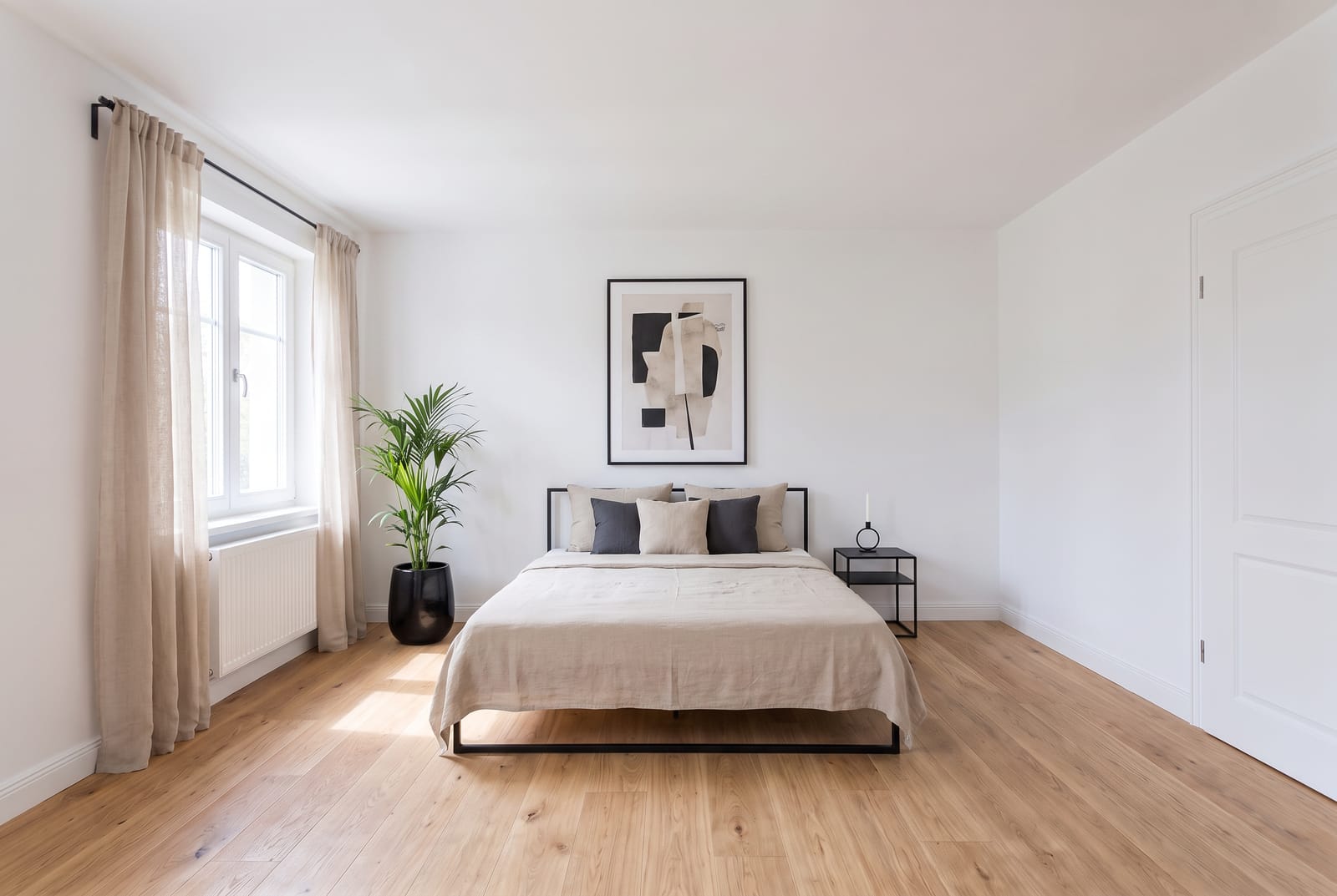 Schlicht eingerichtetes Schlafzimmer mit Bett, zwei Nachttischen, gerahmtem Bild über dem Kopfteil und einer Pflanze neben dem Fenster