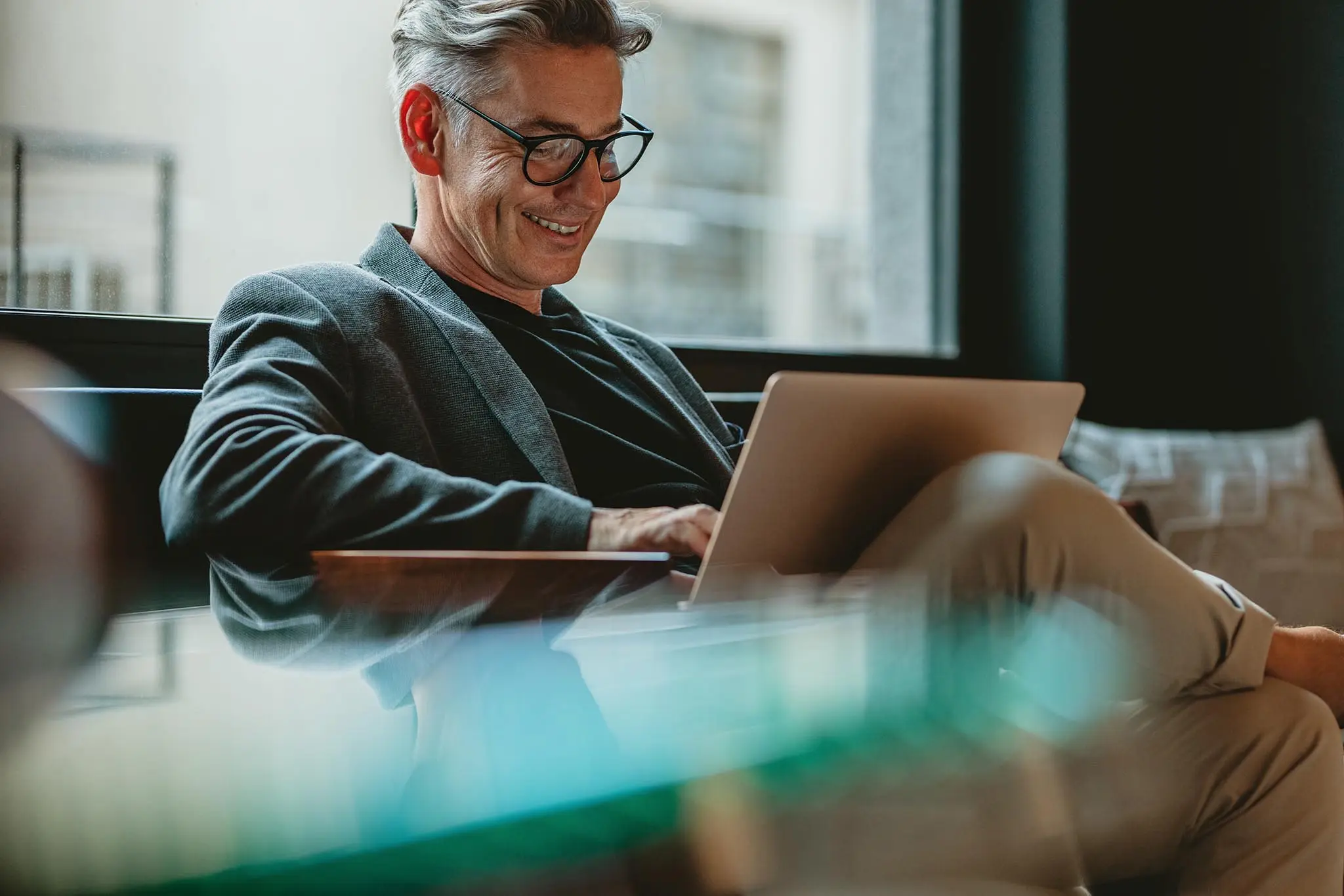 Middle-aged man in glasses working on laptop beside a window with a relaxed posture.