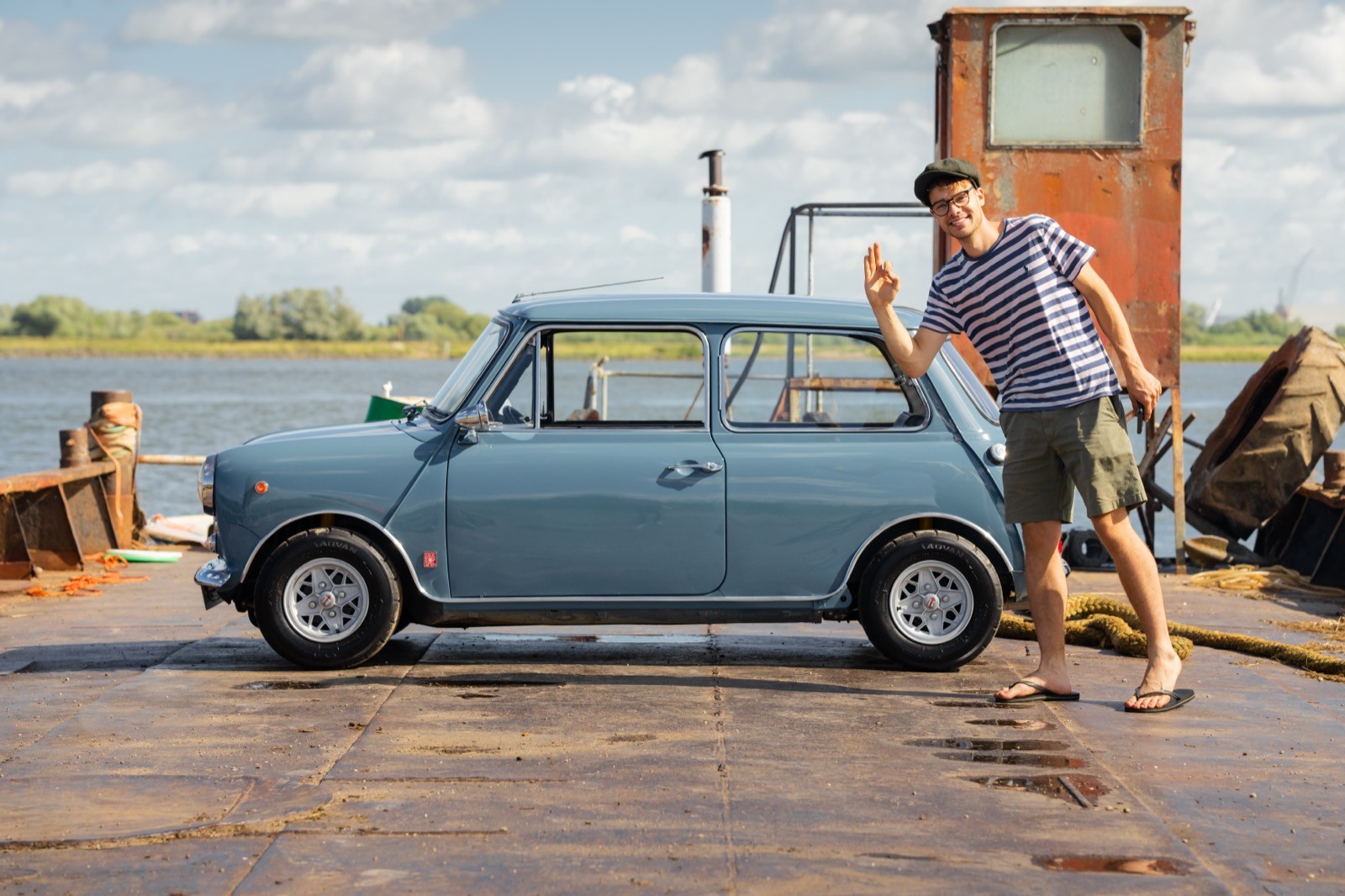 A man leans against a classic blue car parked near water, with a rustic structure in the background under a clear sky.