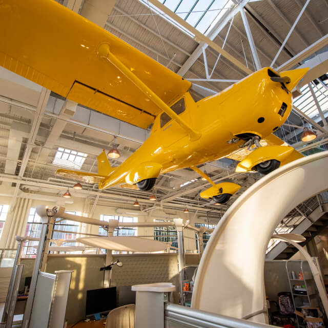 A yellow propeller plane is suspended from the ceiling inside a modern office with cubicles and large windows, reflecting the innovative spirit of this collaborative agency.