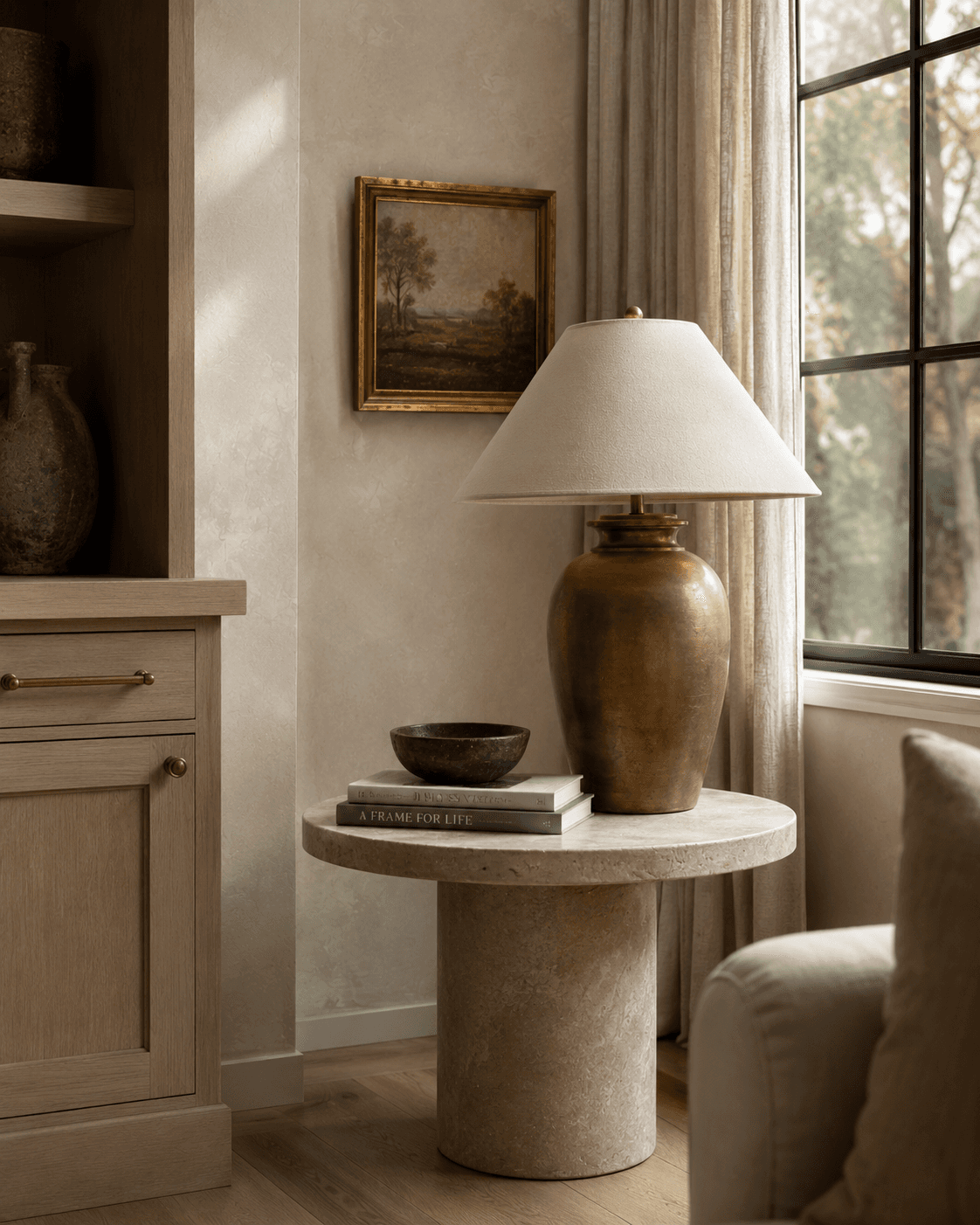 Close-up of a modern French country living room corner with a round limestone side table, aged brass table lamp, stacked books, white oak built-in cabinet, framed landscape artwork, and linen curtains beside a black-framed window.