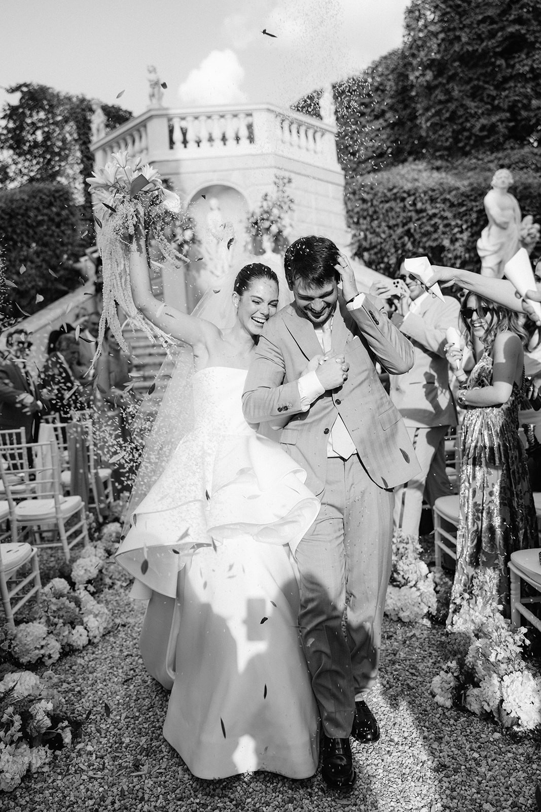 A bride in a white dress and a groom in a black suit hold hands, surrounded by greenery in a romantic setting.