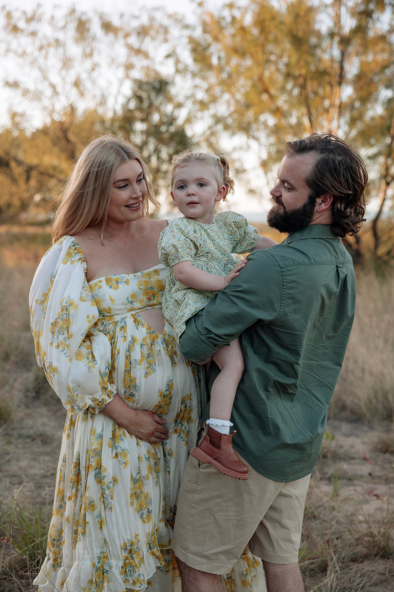 Family maternity session in open grass field in Mackay featuring expectant mother, partner and toddler at golden hour