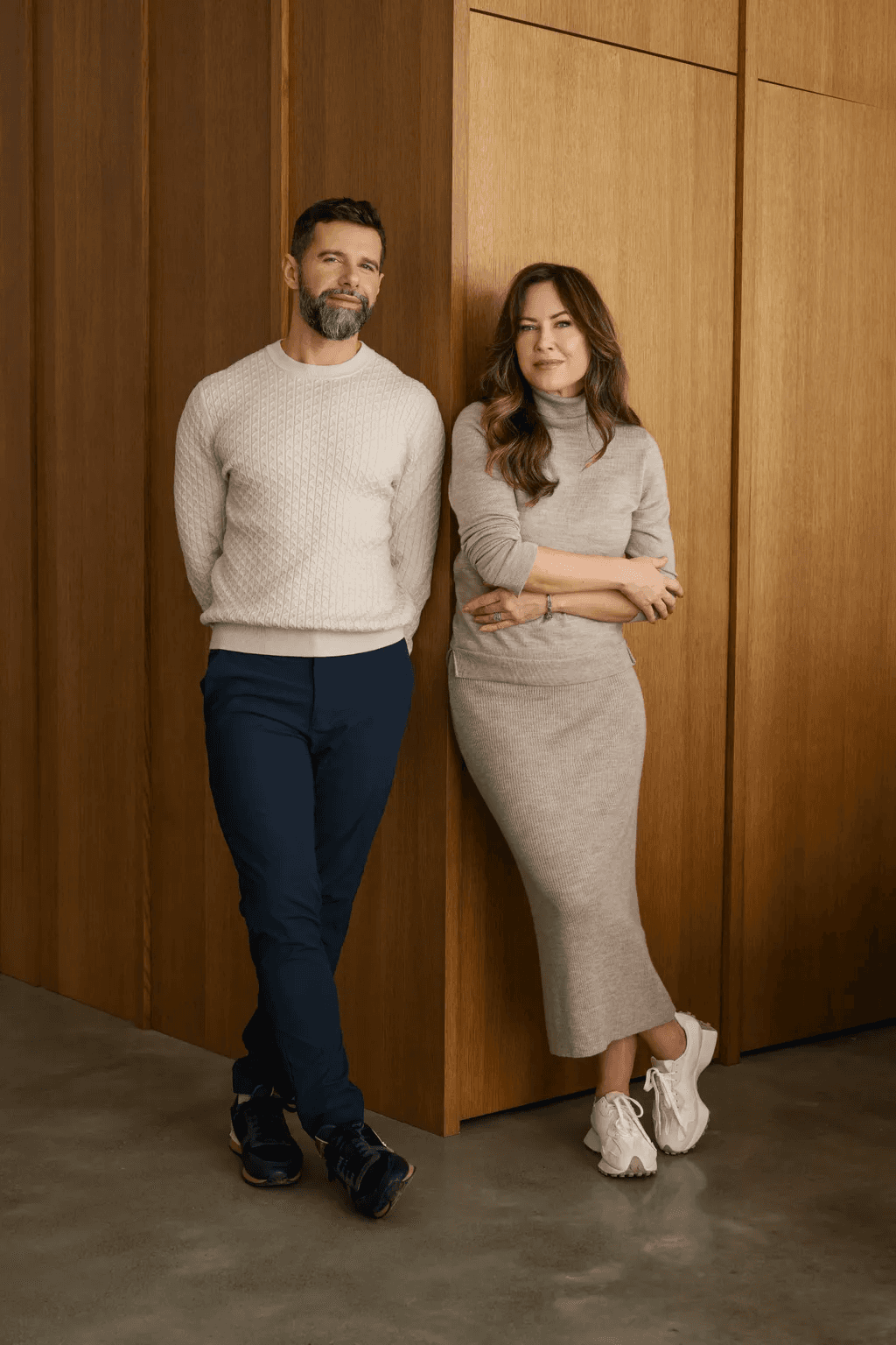A person in a light sweater and dark pants, and a person in a long gray dress with white sneakers, stand casually leaning against a wooden wall in a modern interior.