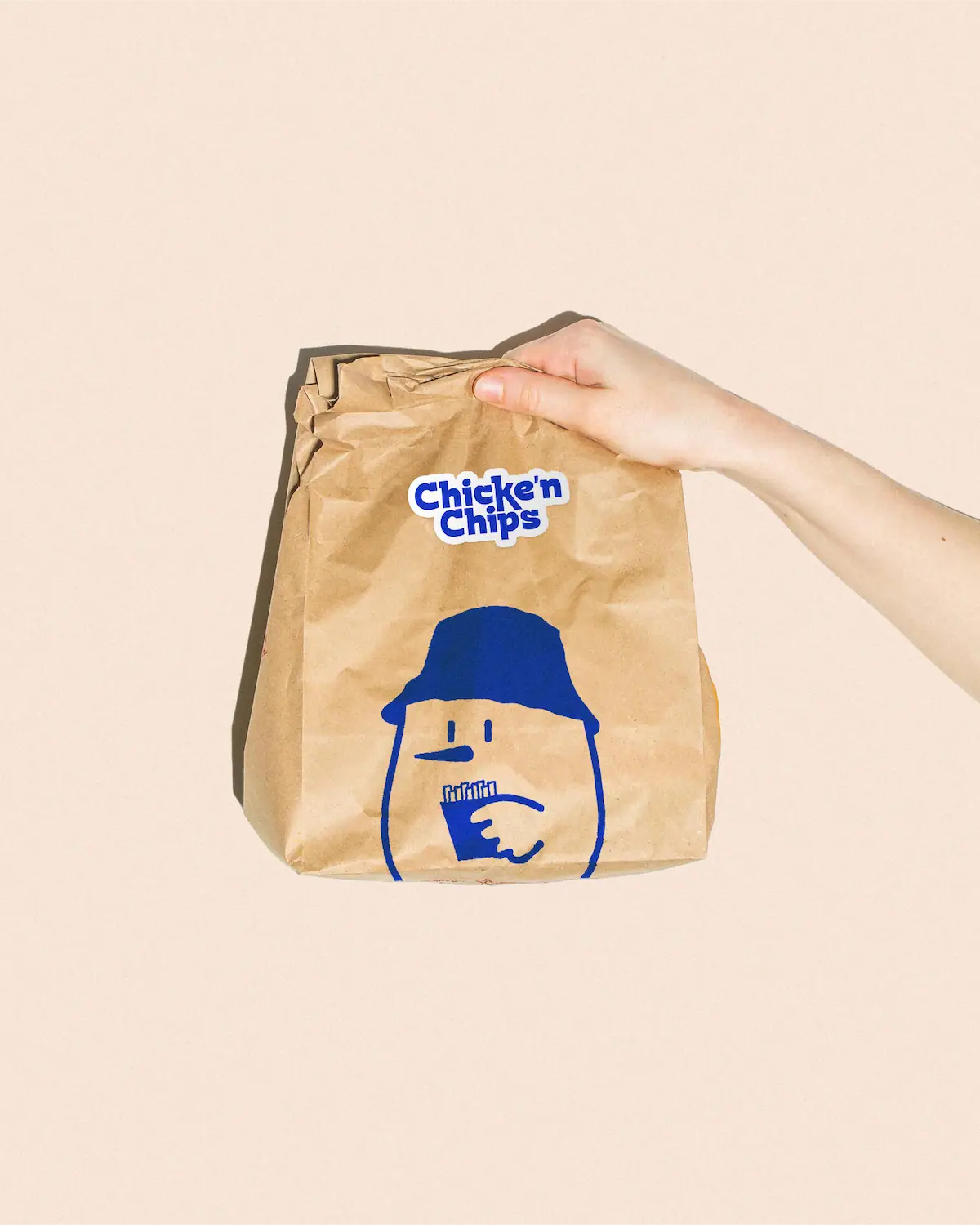 Main tenant un sac kraft de livraison Chicke'n Chips avec la mascotte illustrée en grand format et sticker logo.