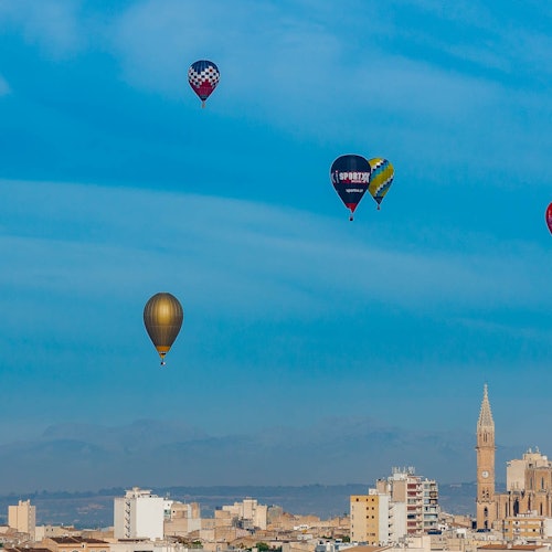 De nombreux montgolfières flottent dans un ciel bleu éclatant au-dessus d'un paysage urbain avec un clocher d'église proéminent.