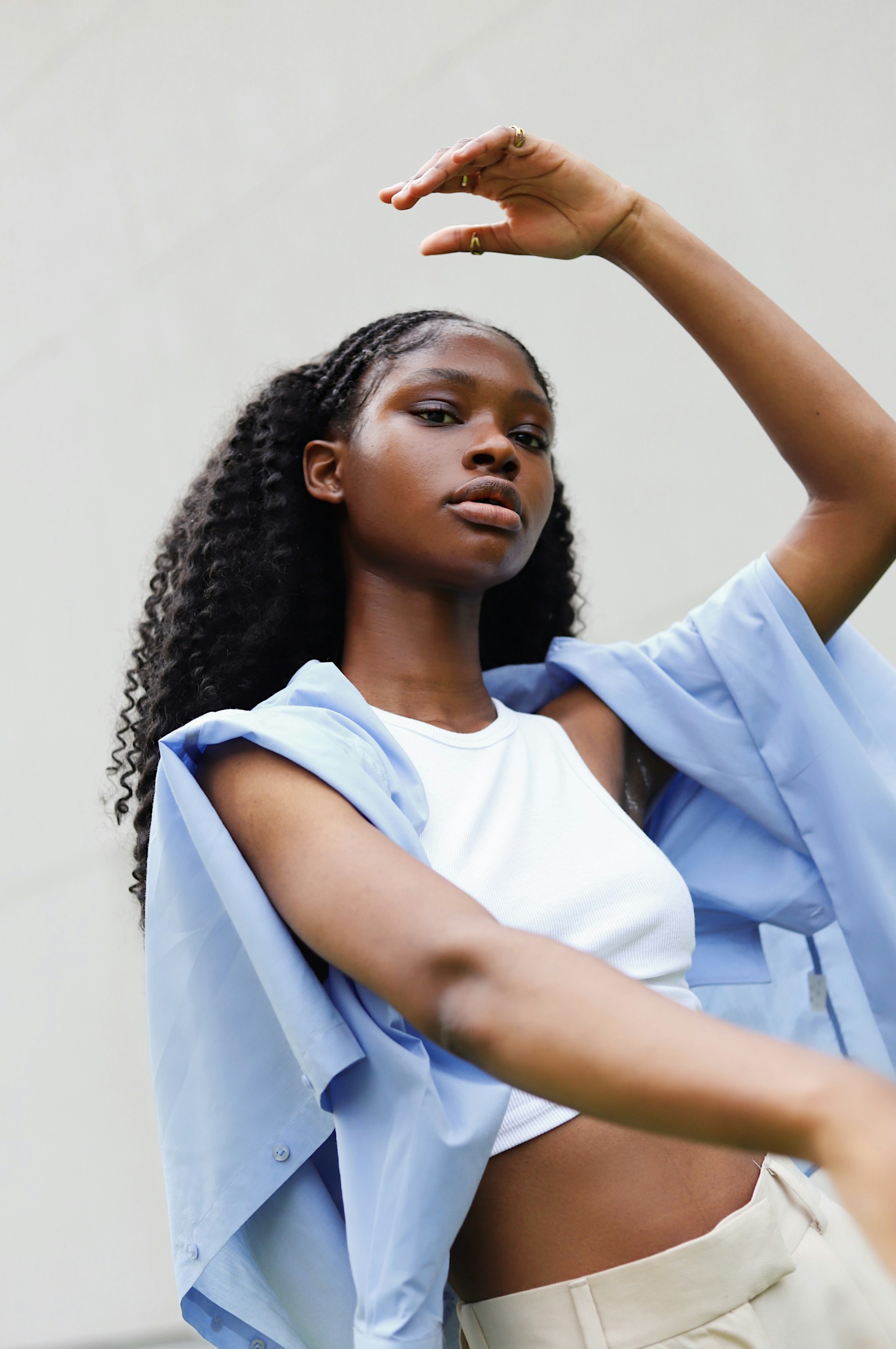 A portrait of a Black woman with long, dark, wavy hair and braids at the top, posing with one arm raised. She is wearing a white crop top, light-colored trousers, and a light blue shirt draped over her shoulders.