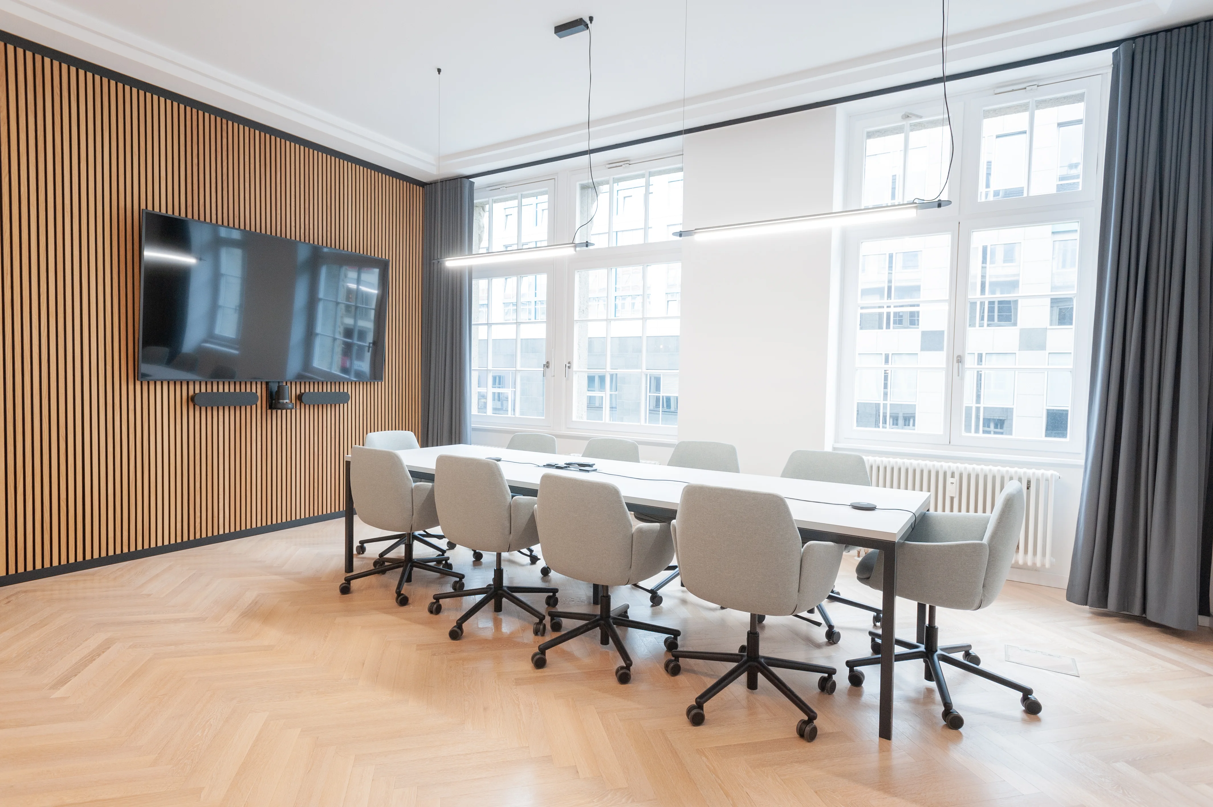 Conference room at AI START-UP BERLIN with a white table, light grey swivel armchairs, a large TV with video bar on a wooden slat wall, grey curtains and herringbone parquet.