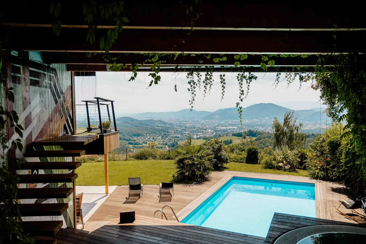 Blick durch Blattwerk auf den Pool, Liegestühle und das weite Tal hinter dem Garten