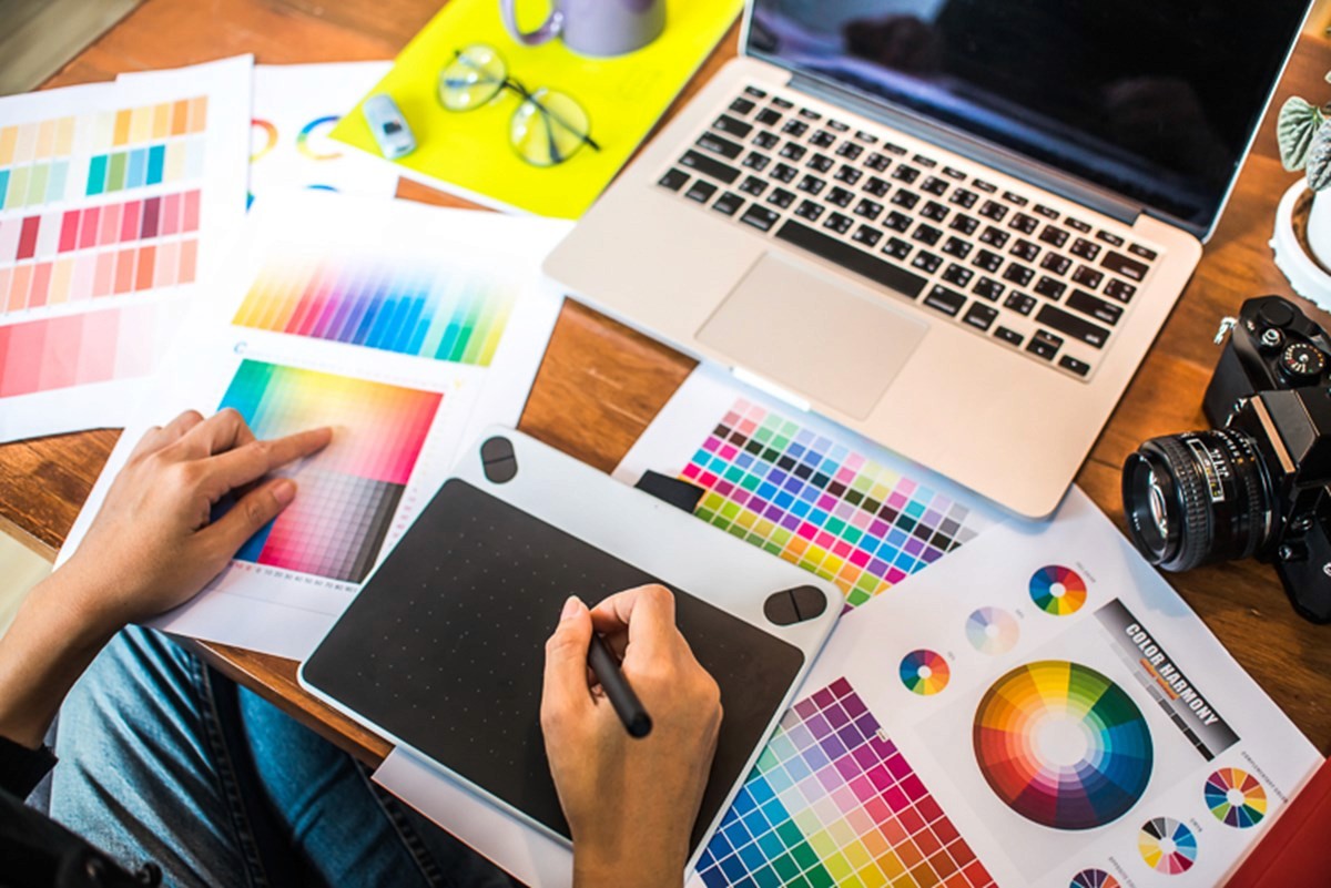 A desk viewed from above, set up for creative design work. Sheets of printed colour palettes and colour wheels are spread across the surface alongside a graphics tablet and stylus in use. A laptop sits at the top of the desk, with a pair of glasses, a camera, and other creative tools nearby. The scene suggests hands‑on work with colour selection and digital design.