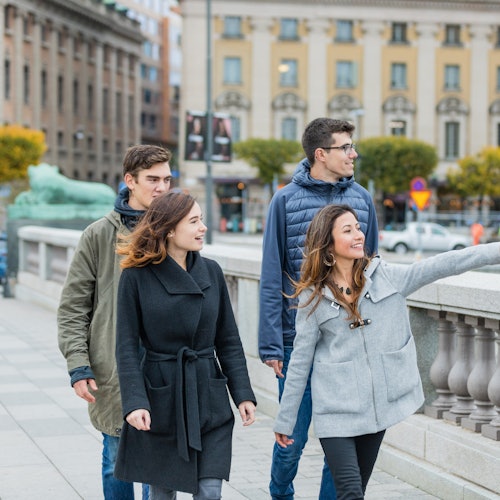 Four people walking and smiling on a city sidewalk, with historic buildings and a street in the background.