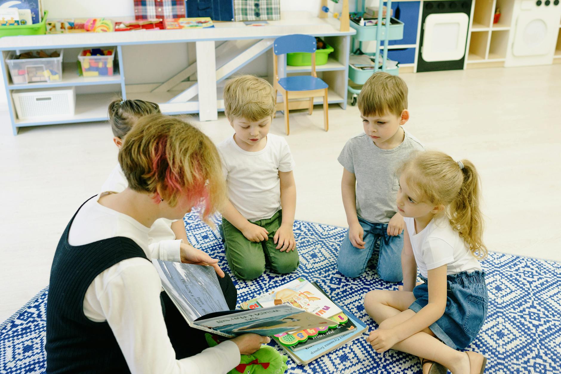 A kindergarten teacher sitting on a low chair reading a colorful picture book to a group of children on a rug.