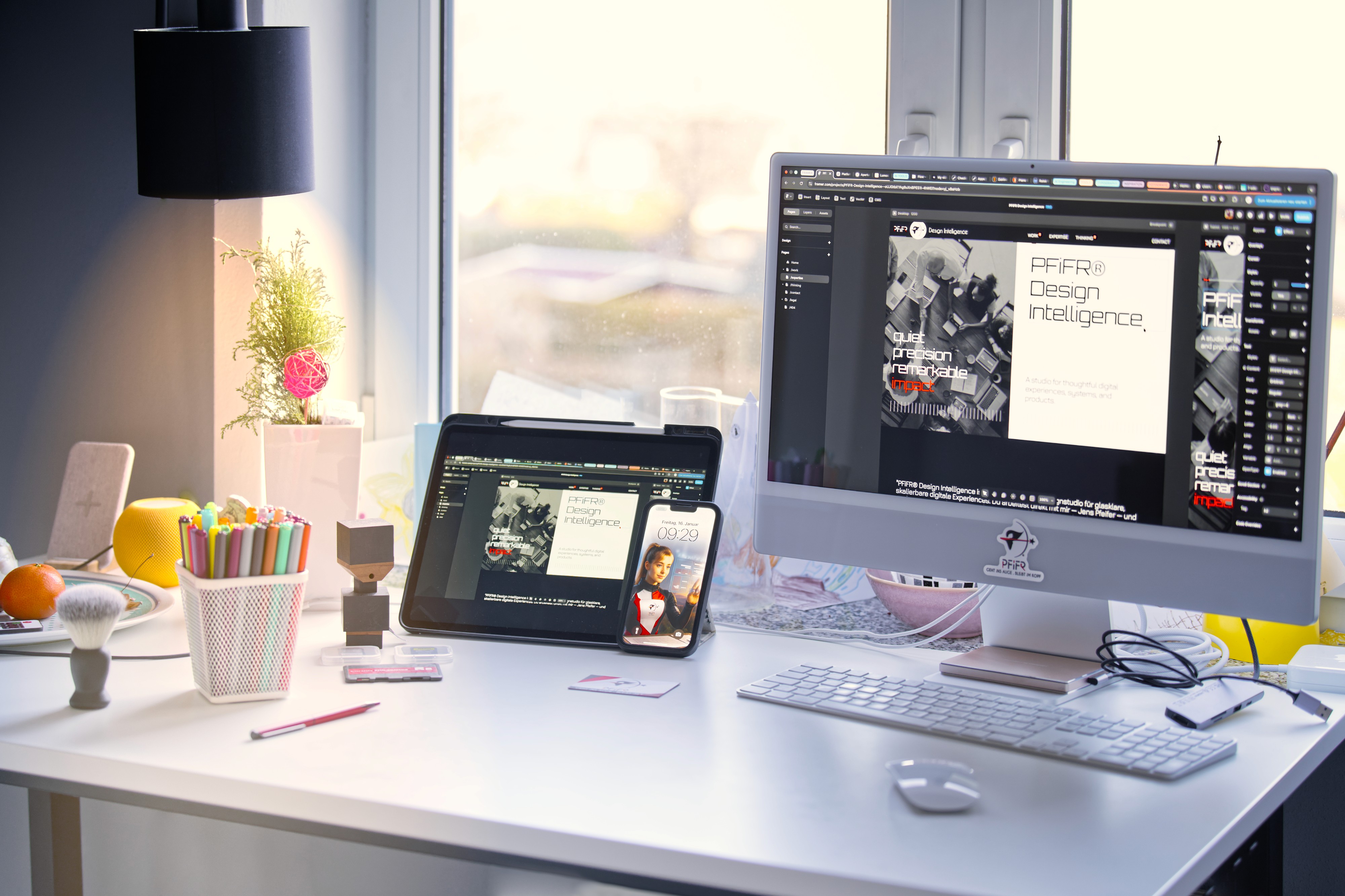 Moderner Design-Workspace mit iMac, iPad und iPhone auf weißem Schreibtisch, geöffnet ist die Website „PFiFR Design Intelligence“, umgeben von Zeichenutensilien, Pflanzen und natürlichem Tageslicht am Fenster.