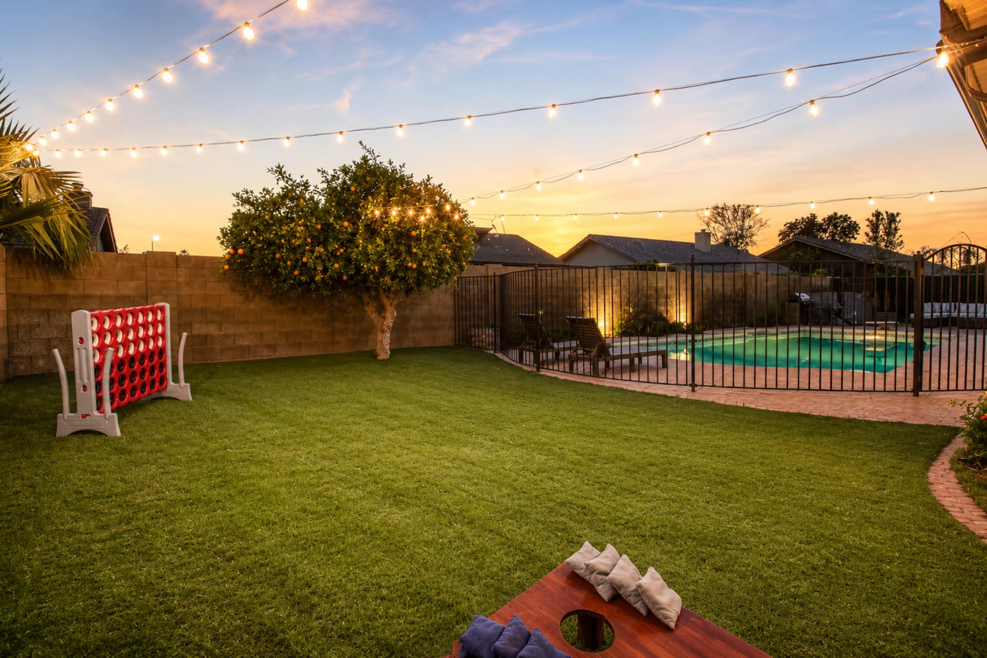 Backyard at sunset with lush turf lawn, pool visible behind wrought iron fence, and string lights overhead. Yard games include giant Connect Four and cornhole. Citrus tree and banana palms.