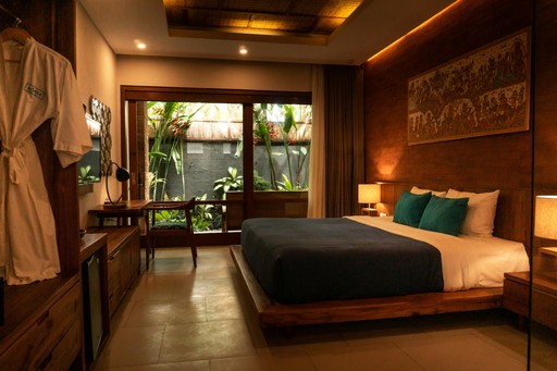 Une chambre d'hôtel chaleureusement éclairée avec des meubles en bois, comprenant un grand lit avec une literie bleue et blanche, deux lampes de chevet, et un bureau face à des portes-fenêtres coulissantes qui s'ouvrent sur un petit jardin tropical à l'extérieur. Un peignoir est suspendu à gauche, et une œuvre d'art décorative est accrochée au-dessus du lit.