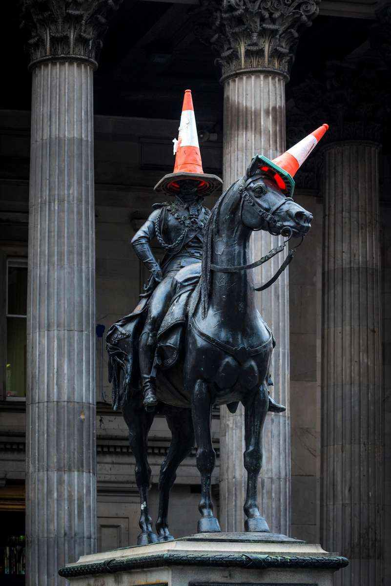 A bronze statue of a man on a horse in front of stone columns, both wearing orange traffic cones on their heads.