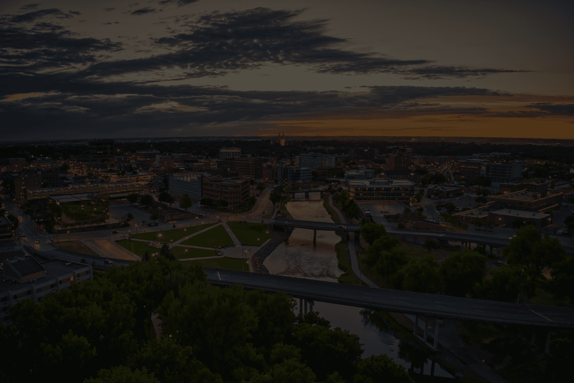 Aerial view of a city at sunset with buildings, bridges, and a river running through the center.