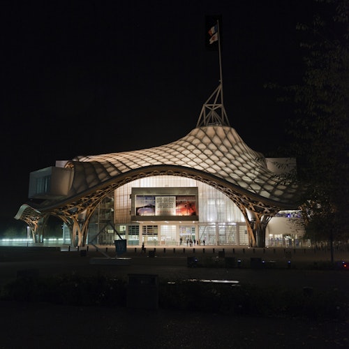 一座现代建筑，屋顶呈独特波浪状，夜间灯火通明，顶部飘扬着一面旗帜，右侧部分树木隐约可见。