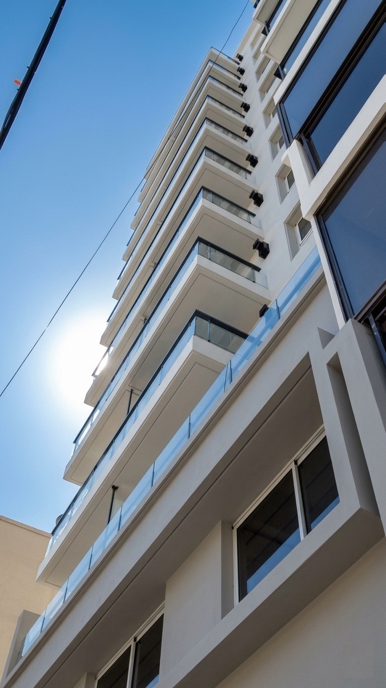 Vista en contrapicado de edificio de departamentos con revestimiento blanco y balcones con baranda de vidrio, BA Homes, Rosario