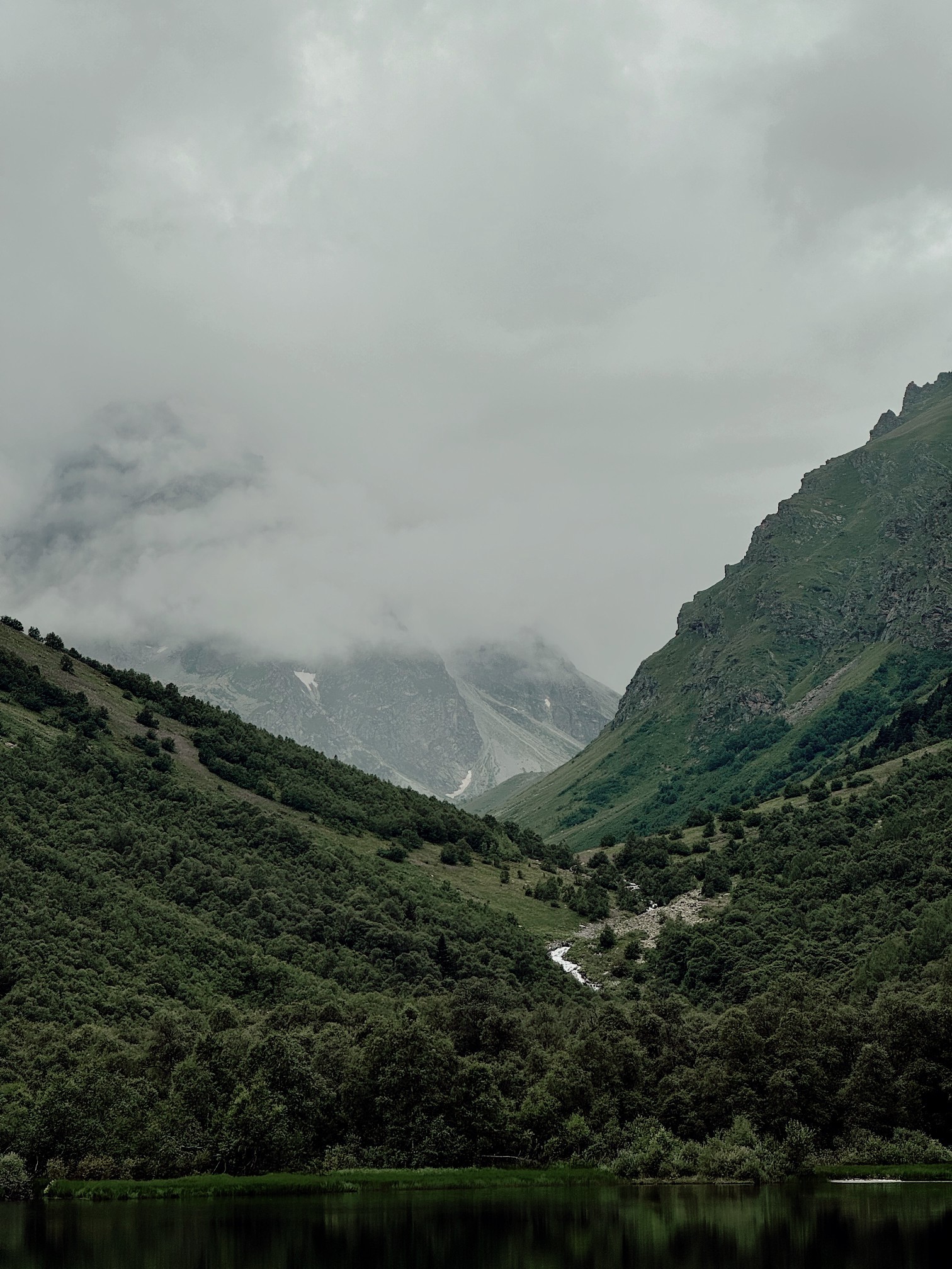 A narrow green mountain valley with steep hillsides, a calm dark lake in the foreground, and heavy clouds hanging low over the distant peaks.