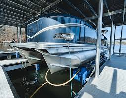 A sleek, modern pontoon boat with a blue and silver metallic finish is docked in a covered marina, reflecting sunlight on the water surface.