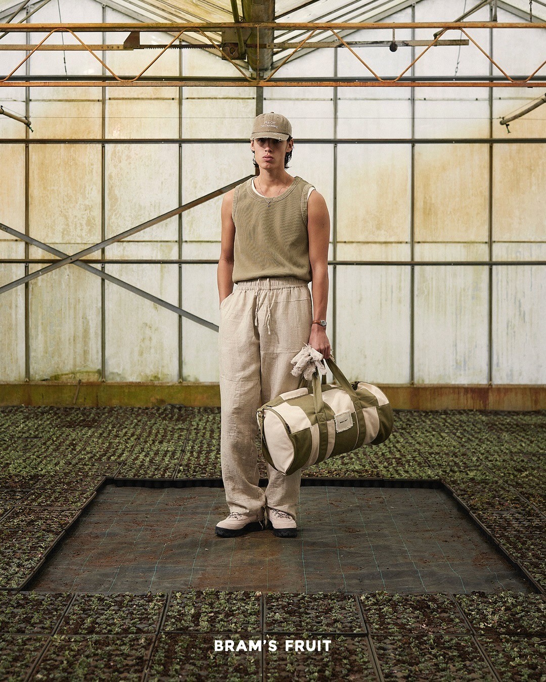 Extrait du lookbook Brams Fruit, le mannequin dans une serre porte un ensemble en camieu de beige/vert, pantalon, débardeur, casquette et sac de sport