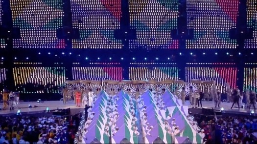 The image depicts a vibrant stage performance featuring dancers in colorful costumes, pyramidal structures adorned with stripes and floral patterns, and a dazzling backdrop of multicolored LED lights.
