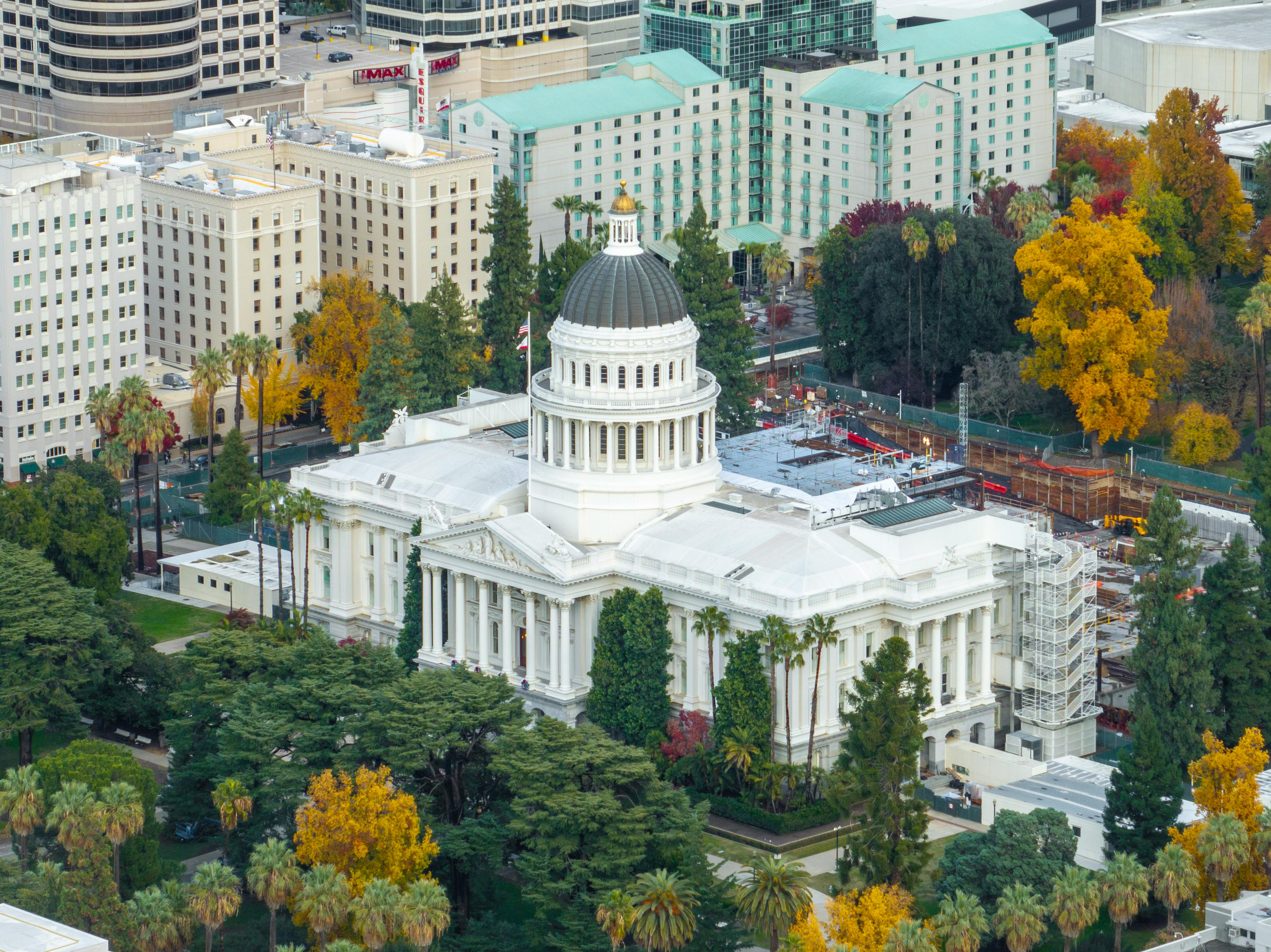 Aerial view of the California State Capitol building with green trees and fall foliage surrounding it.
