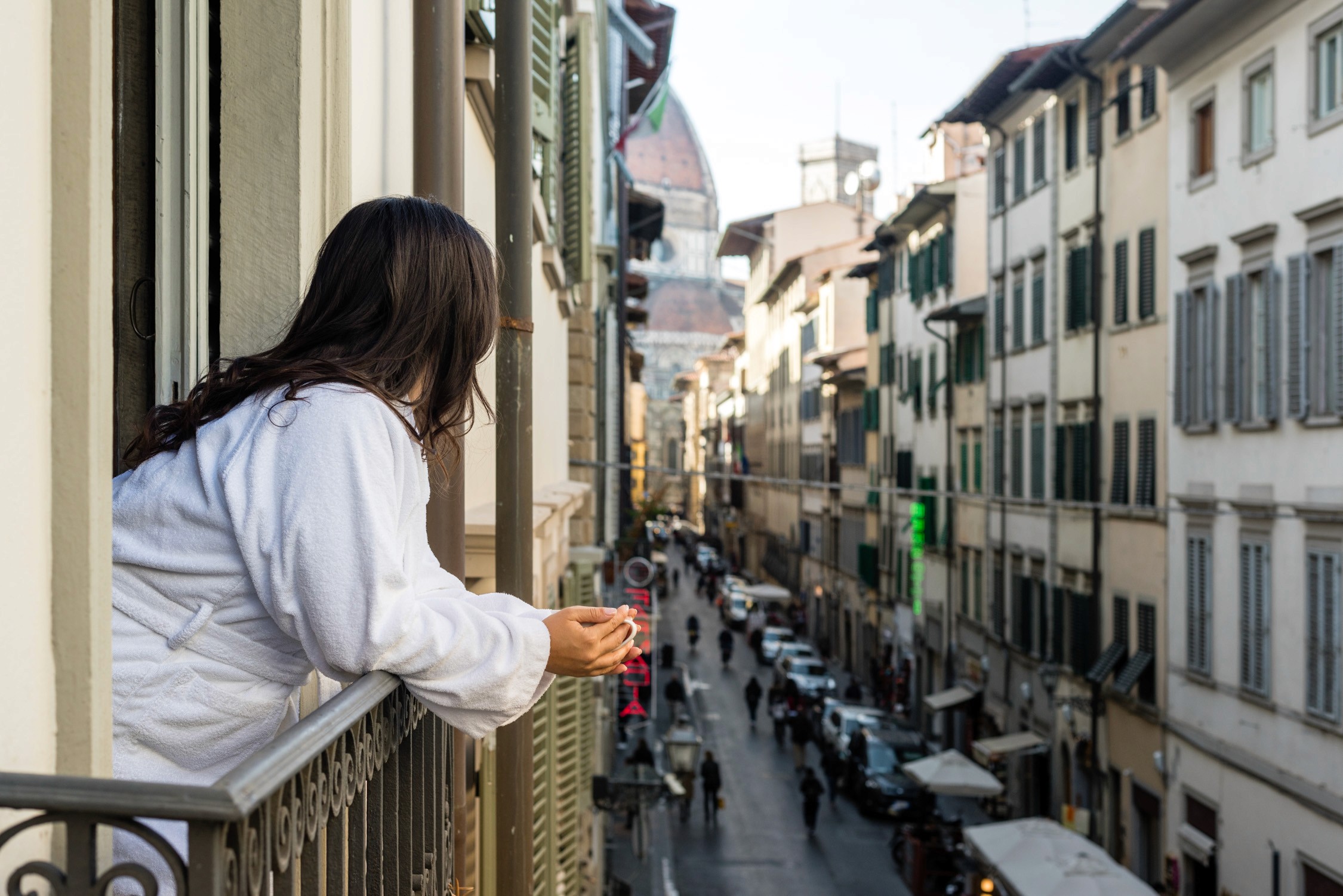 A woman in a white sweater leans on a balcony railing overlooking a narrow European street with shops and cafes.